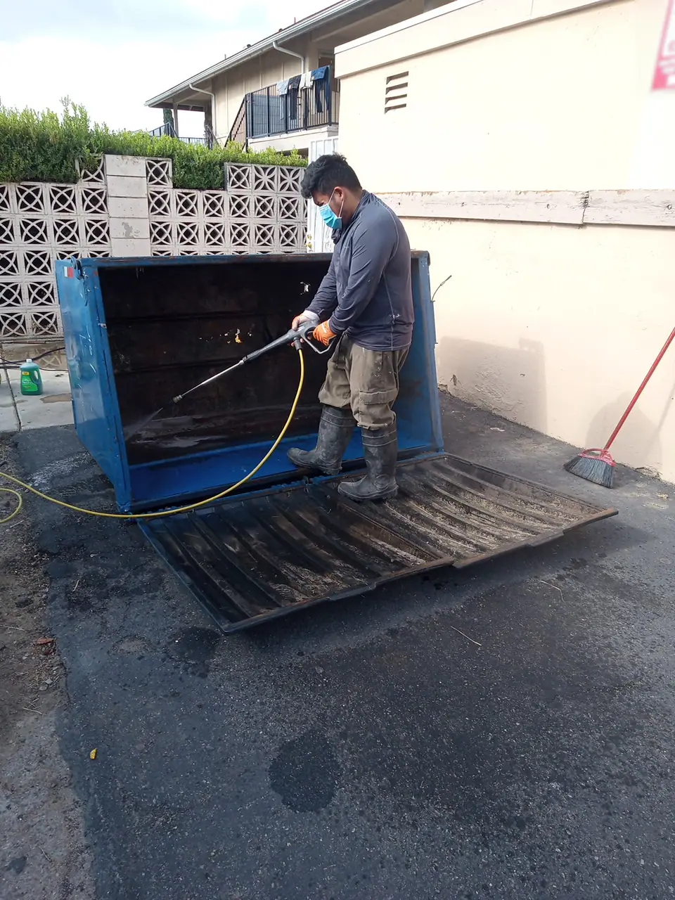 Crew member in a mask washing out an empty dumpster bin behind an apartment building