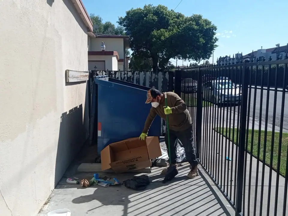 Crew member picking up boxes and trash around a dumpster in an apartment enclosure