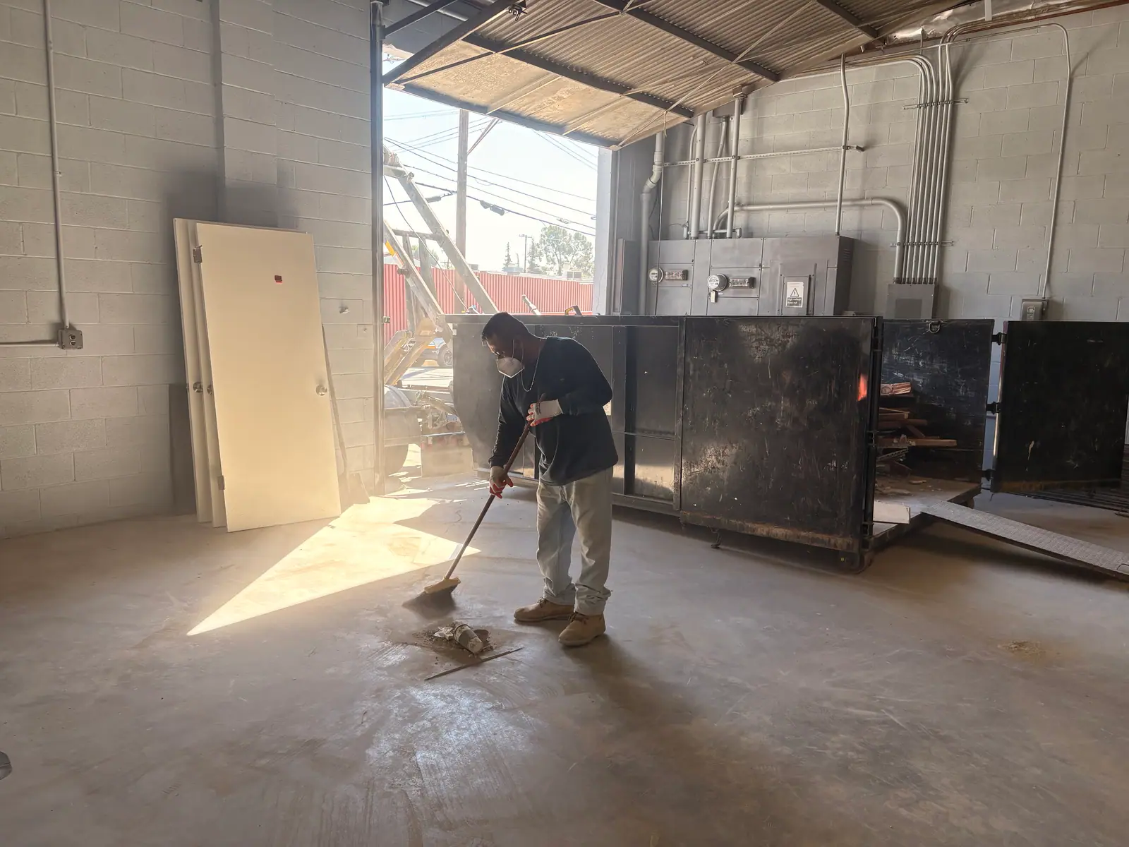 Crew member in a mask sweeping a warehouse floor next to an open roll-off bin